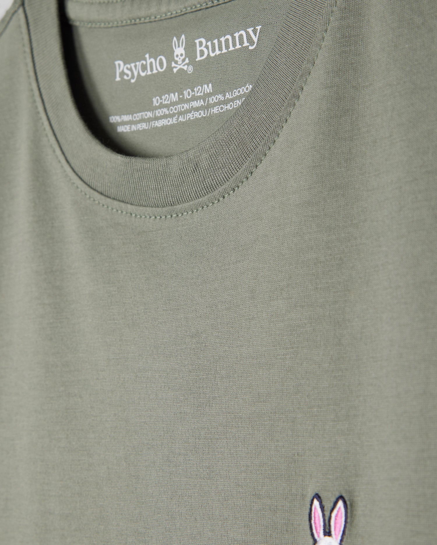 A green KIDS CLASSIC CREW NECK TEE - B0U014B200 by Psycho Bunny features a pink and white embroidered bunny skull logo on the lower side. Made from 100% Pima cotton, it's ideal for back-to-school outfits, with size info in white text on the inner back neck.