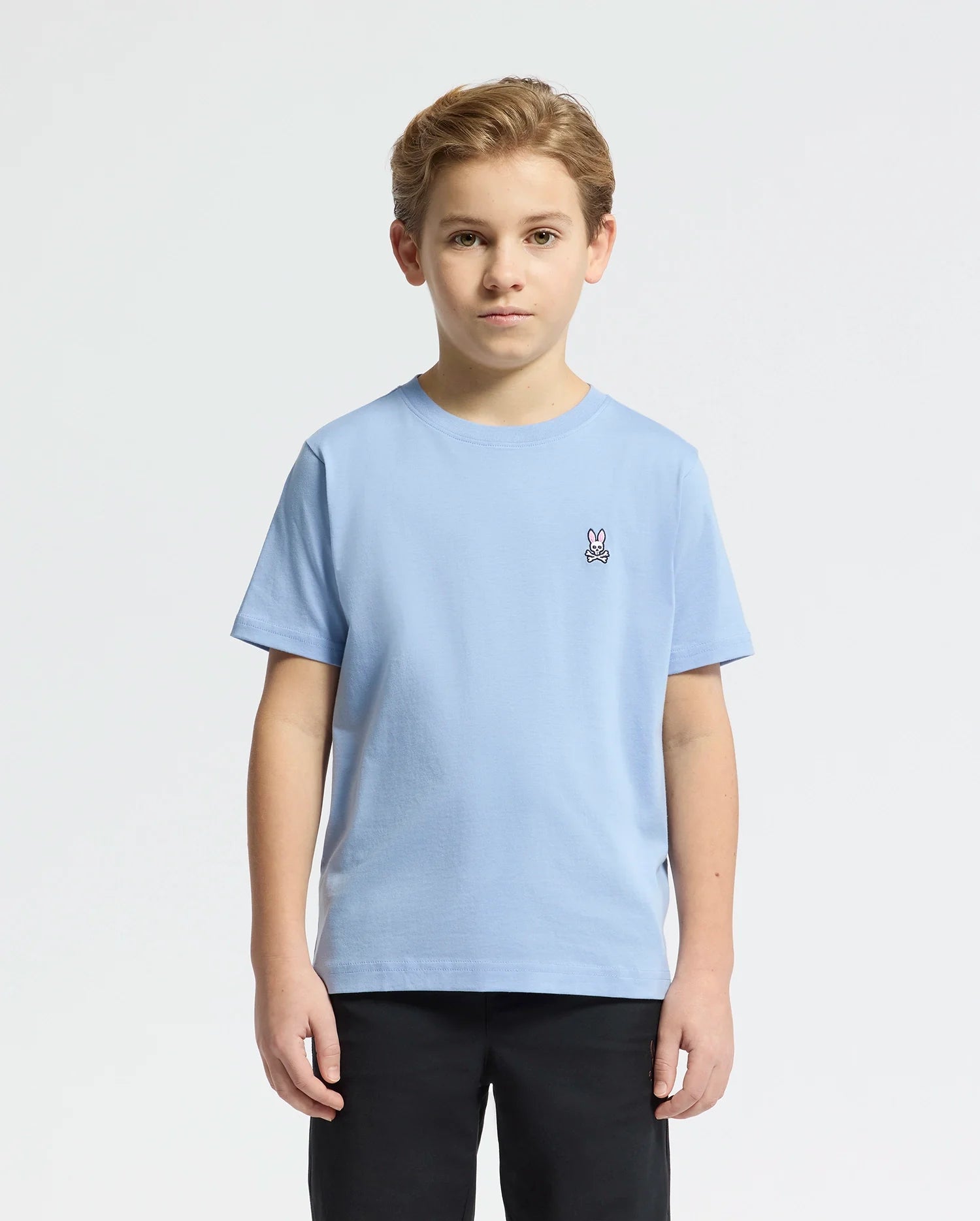 A young boy wears the Psycho Bunny KIDS CLASSIC CREW NECK TEE (B0U014B200) in light blue Pima cotton with an embroidered bunny logo on the upper left. He stands against a plain white background, facing the camera.
