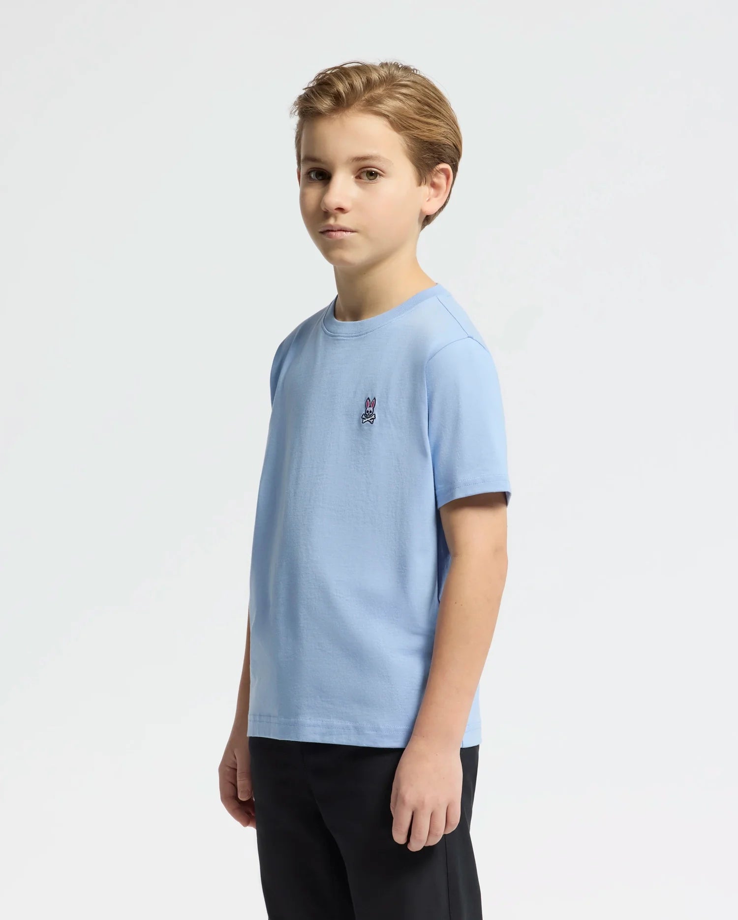 A young boy with short light brown hair stands against a plain white background, wearing the KIDS CLASSIC CREW NECK TEE (B0U014B200) by Psycho Bunny. The light blue tee is made of soft Pima cotton, features a small Bunny logo on the chest, and is paired with dark pants. His gaze is steady and direct.