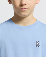 A person wears the Kids Classic Crew Neck Tee by Psycho Bunny, crafted from soft Pima cotton in light blue, featuring a subtle embroidered Bunny logo with crossbones. The focus is on the upper half of the tee and part of their face against a plain background.