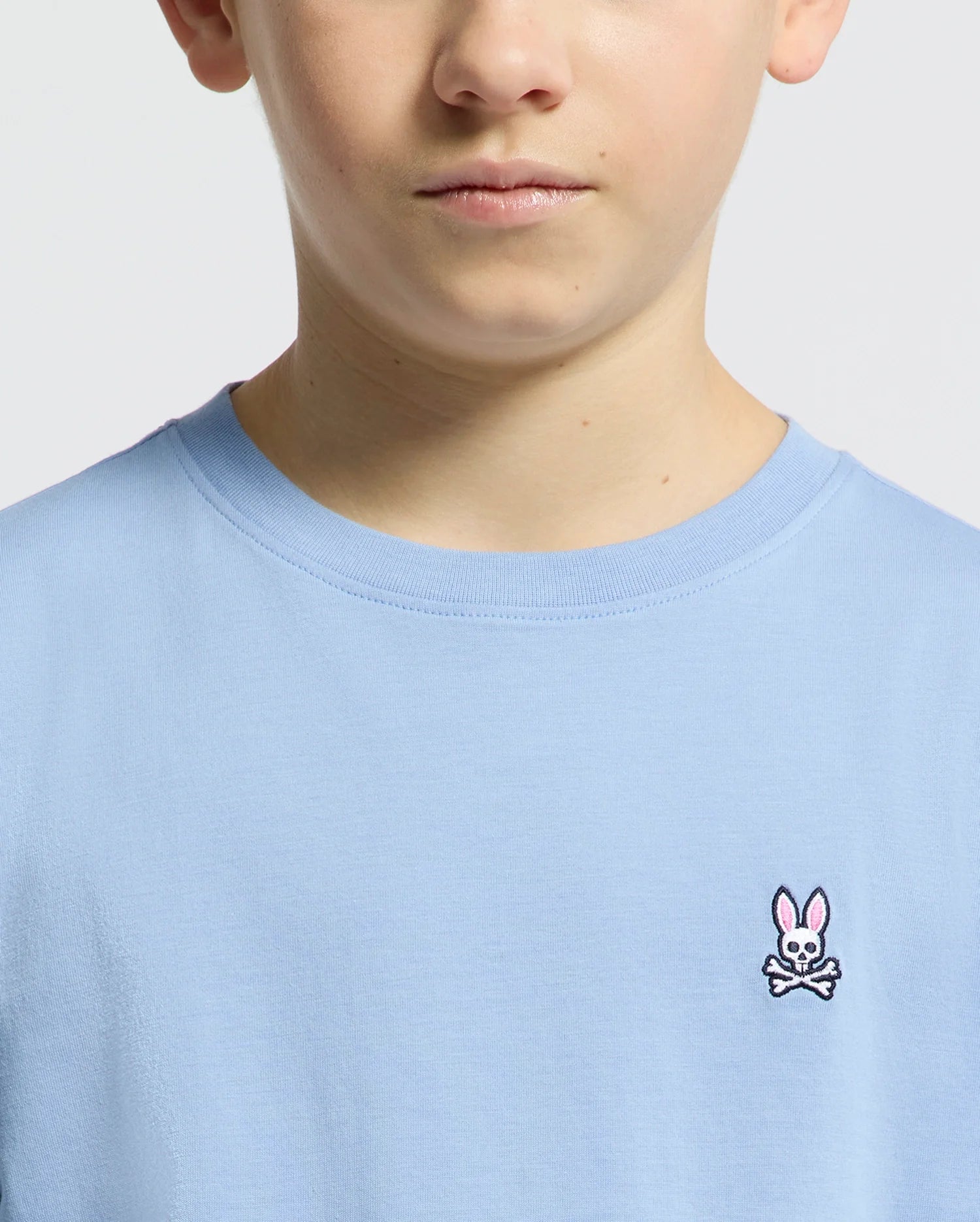 A person wears the Kids Classic Crew Neck Tee by Psycho Bunny, crafted from soft Pima cotton in light blue, featuring a subtle embroidered Bunny logo with crossbones. The focus is on the upper half of the tee and part of their face against a plain background.
