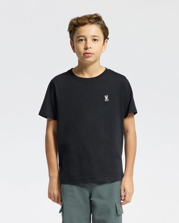 A young person with brown hair is wearing a Psycho Bunny KIDS CLASSIC CREW NECK TEE, made of 100% Pima cotton, showing a small white skull and bunny logo on the left chest. They pair it with green cargo pants against a plain white background.