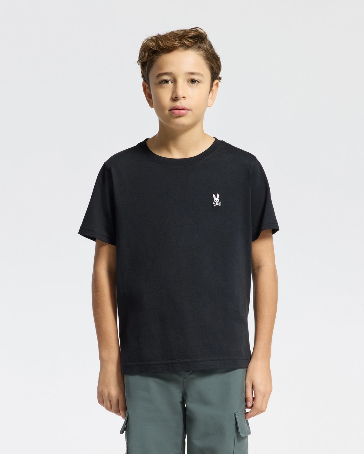 A young person with brown hair is wearing a Psycho Bunny KIDS CLASSIC CREW NECK TEE, made of 100% Pima cotton, showing a small white skull and bunny logo on the left chest. They pair it with green cargo pants against a plain white background.