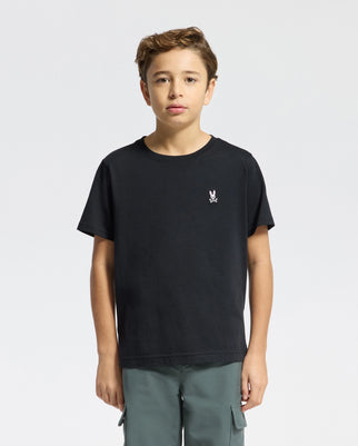 A young person with brown hair is wearing a Psycho Bunny KIDS CLASSIC CREW NECK TEE, made of 100% Pima cotton, showing a small white skull and bunny logo on the left chest. They pair it with green cargo pants against a plain white background.