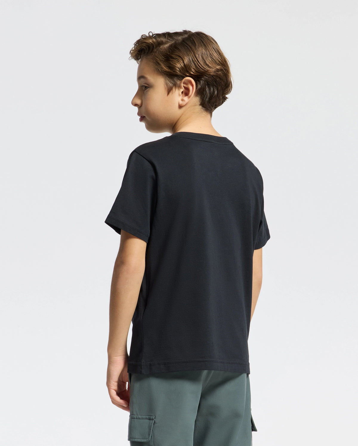 A young boy with short hair stands sideways, sporting a Psycho Bunny KIDS CLASSIC CREW NECK TEE - B0U014CRPC in black and green cargo shorts against a plain white background.