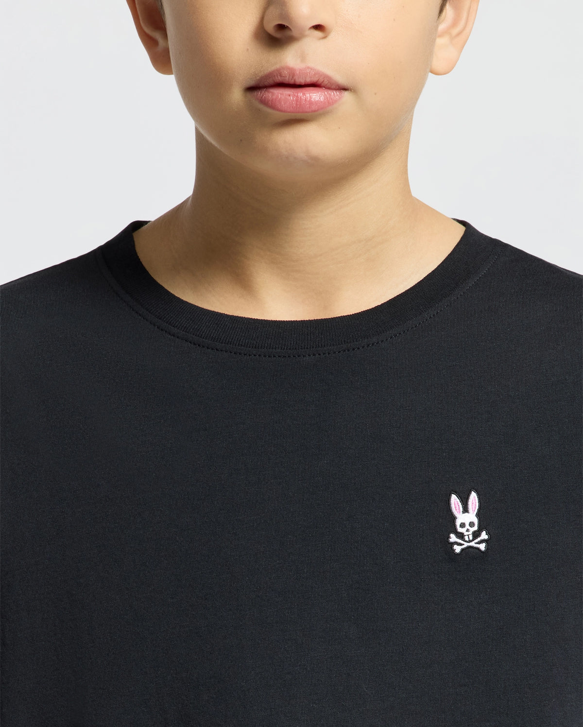 A person wearing the Psycho Bunny KIDS CLASSIC CREW NECK TEE - B0U014CRPC in classic black, made from 100% Pima cotton, features a chest logo with a pink-eared rabbit and crossbones on a plain light-colored background.