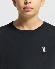 A person wearing the Psycho Bunny KIDS CLASSIC CREW NECK TEE - B0U014CRPC in classic black, made from 100% Pima cotton, features a chest logo with a pink-eared rabbit and crossbones on a plain light-colored background.
