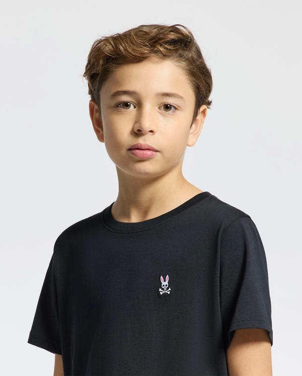 A child with short light brown hair stands against a plain background, wearing Psycho Bunny's KIDS CLASSIC CREW NECK TEE in black. This stylish tee showcases a playful rabbit and crossbones design on the chest and is crafted from 100% Pima cotton for ultimate comfort.