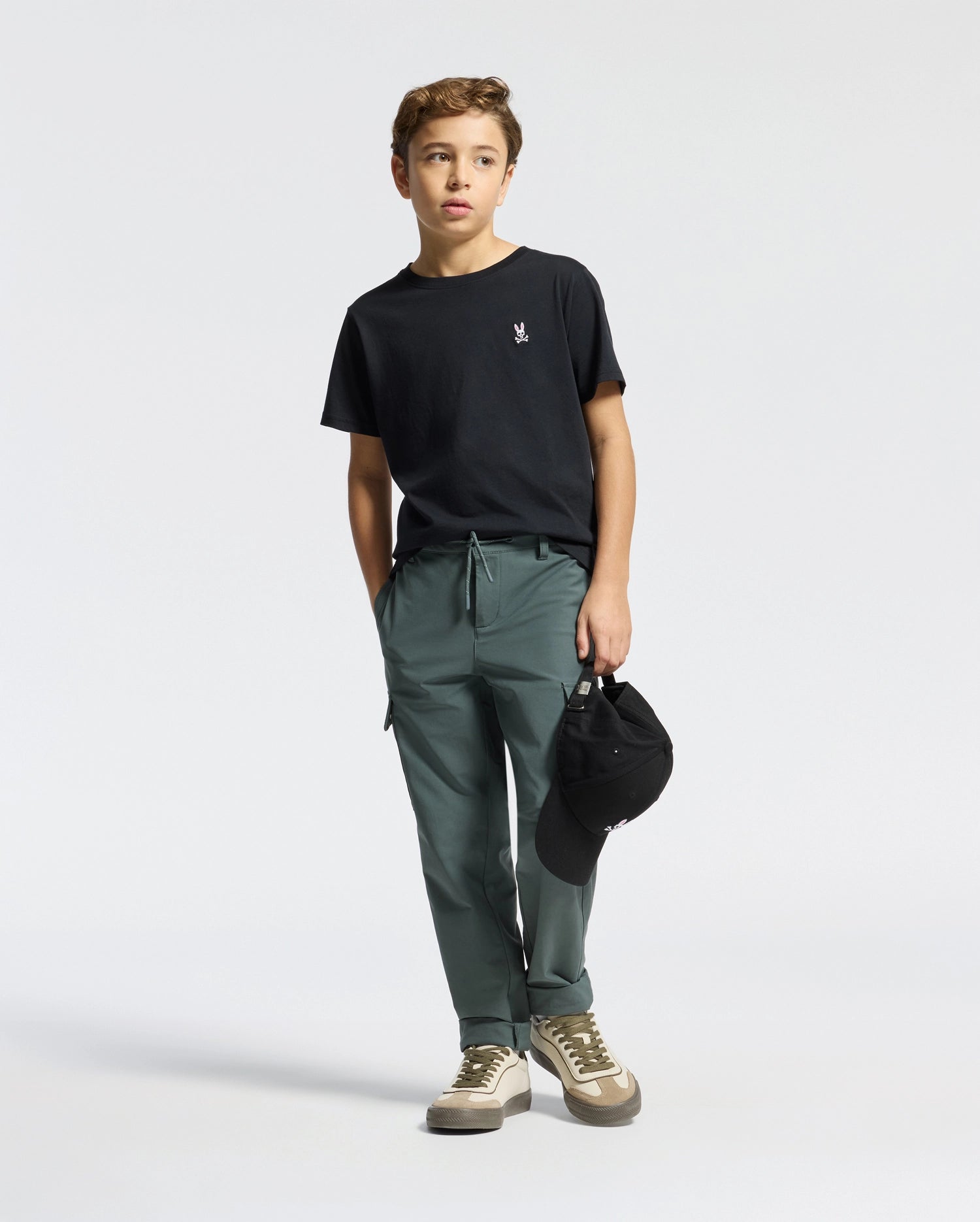 A young boy stands against a plain background, wearing a Psycho Bunny KIDS CLASSIC CREW NECK TEE - B0U014CRPC. His black tee is 100% Pima cotton and pairs with green pants and sneakers. Holding a black cap in one hand, he gazes slightly to the side.
