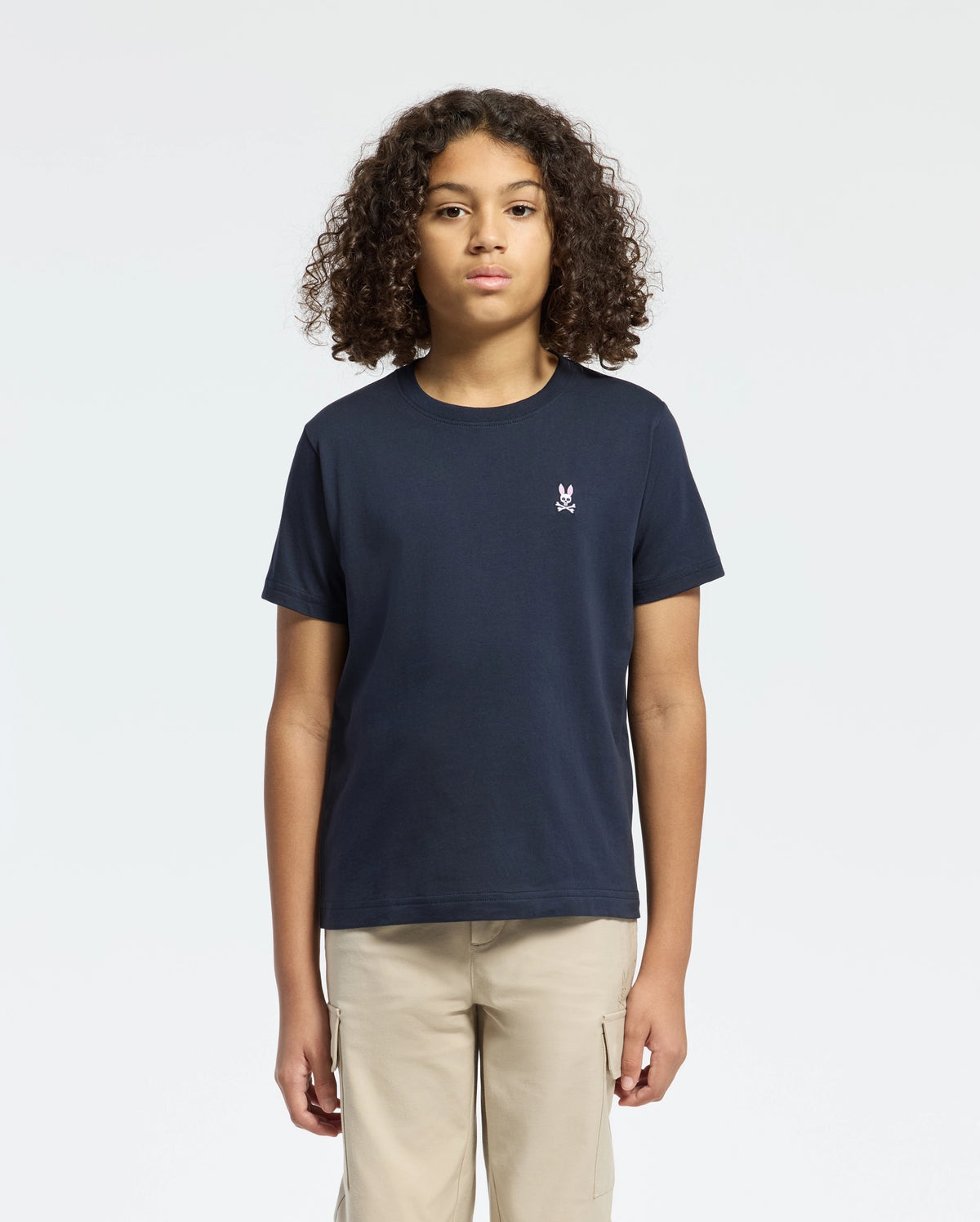 A curly-haired person wears a Psycho Bunny Kids Classic Crew Neck Tee in plain navy, made from 100% Pima cotton with a small chest embroidery. Paired with beige pants against a white background, they represent timeless basics.