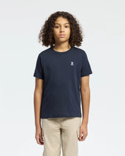 A curly-haired person wears a Psycho Bunny Kids Classic Crew Neck Tee in plain navy, made from 100% Pima cotton with a small chest embroidery. Paired with beige pants against a white background, they represent timeless basics.