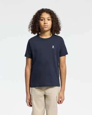 A curly-haired person wears a Psycho Bunny Kids Classic Crew Neck Tee in plain navy, made from 100% Pima cotton with a small chest embroidery. Paired with beige pants against a white background, they represent timeless basics.