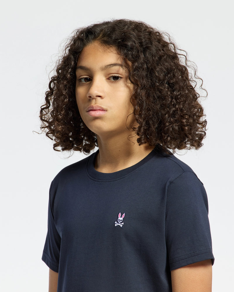 A curly-haired person wears a navy Psycho Bunny KIDS CLASSIC CREW NECK TEE - B0U014CRPC, made from 100% Pima cotton, showcasing a small pink bunny with crossbones on the chest, standing out against the white background.