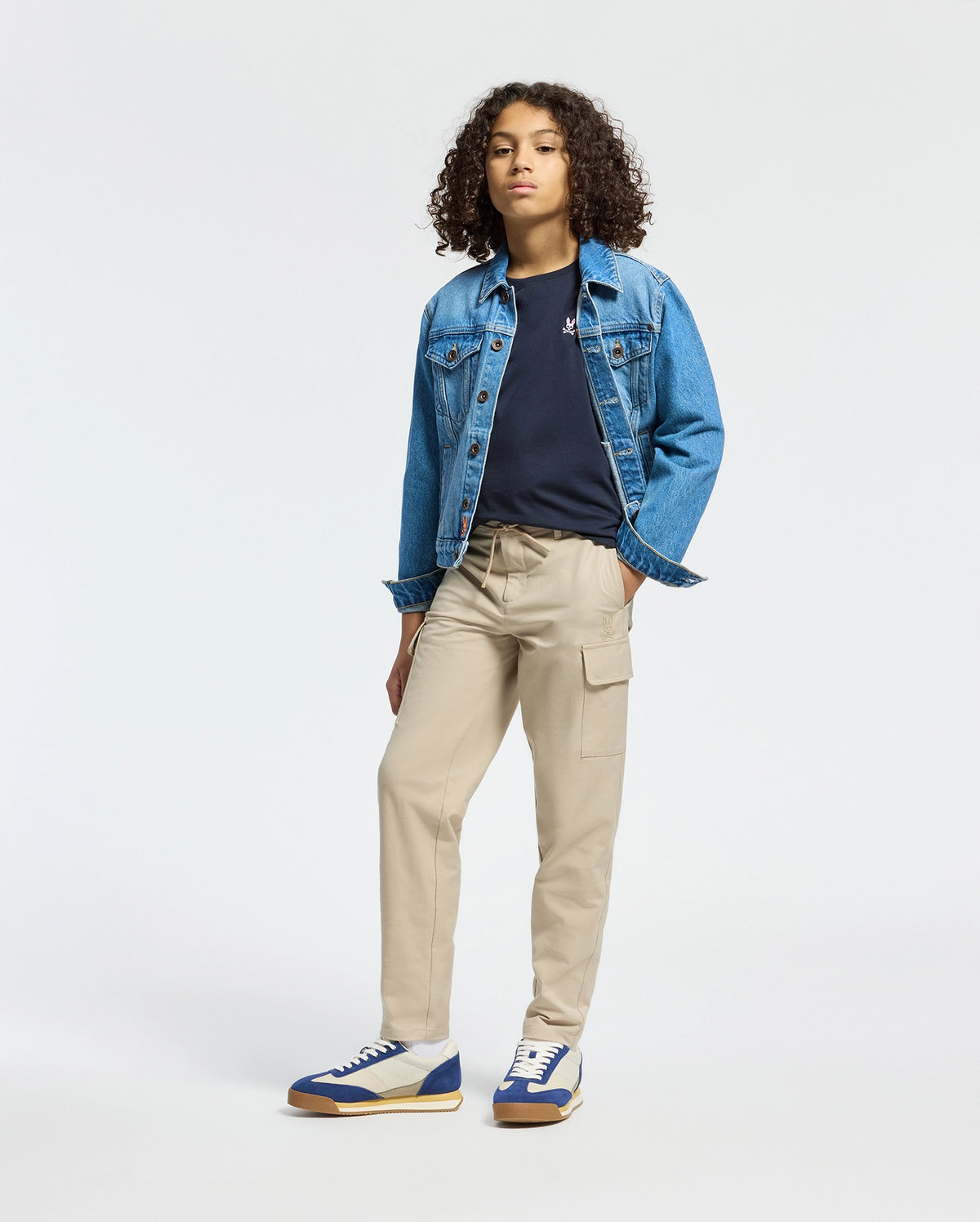 A person with curly hair wearing a classic denim jacket, a Psycho Bunny KIDS CLASSIC CREW NECK TEE in black, beige cargo pants, and blue and white sneakers stands confidently against a plain white background.