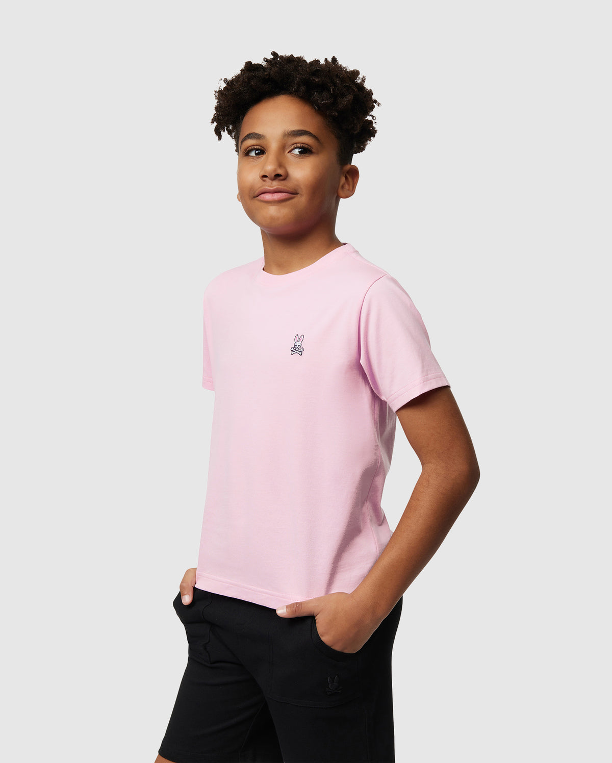A young person with short curly hair wears Psycho Bunny's KIDS CLASSIC CREW NECK TEE in light pink, made from 100% Pima cotton, featuring a small bunny logo and paired with black shorts. They stand against a white background, hands in pockets and slightly smiling—ideal for back-to-school outfits.
