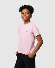 A young person with short curly hair wears Psycho Bunny's KIDS CLASSIC CREW NECK TEE in light pink, made from 100% Pima cotton, featuring a small bunny logo and paired with black shorts. They stand against a white background, hands in pockets and slightly smiling—ideal for back-to-school outfits.