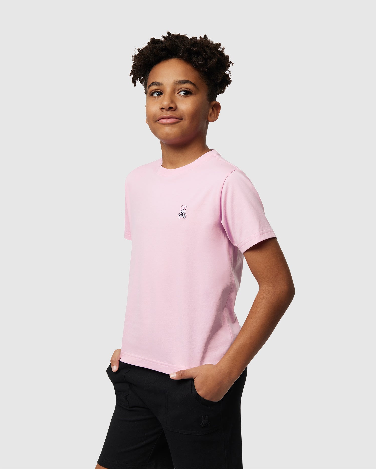 A young person with short curly hair wears Psycho Bunny's KIDS CLASSIC CREW NECK TEE in light pink, made from 100% Pima cotton, featuring a small bunny logo and paired with black shorts. They stand against a white background, hands in pockets and slightly smiling—ideal for back-to-school outfits.