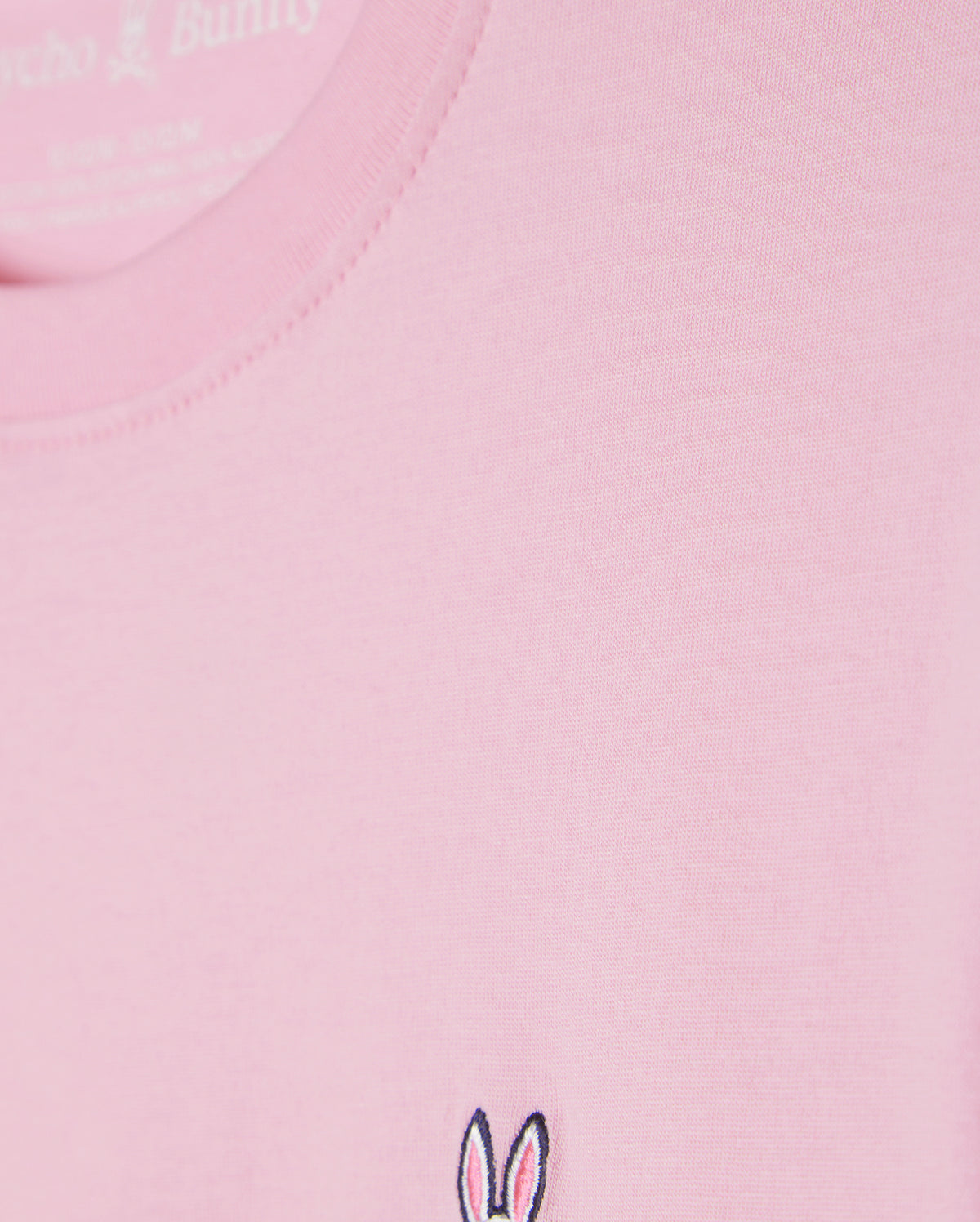 Close-up of the KIDS CLASSIC CREW NECK TEE - B0U014CRPC from Psycho Bunny, a light pink shirt made from 100% Pima cotton. It features a small embroidered bunny ears and skull logo near the hem, ideal for stylish back-to-school outfits.