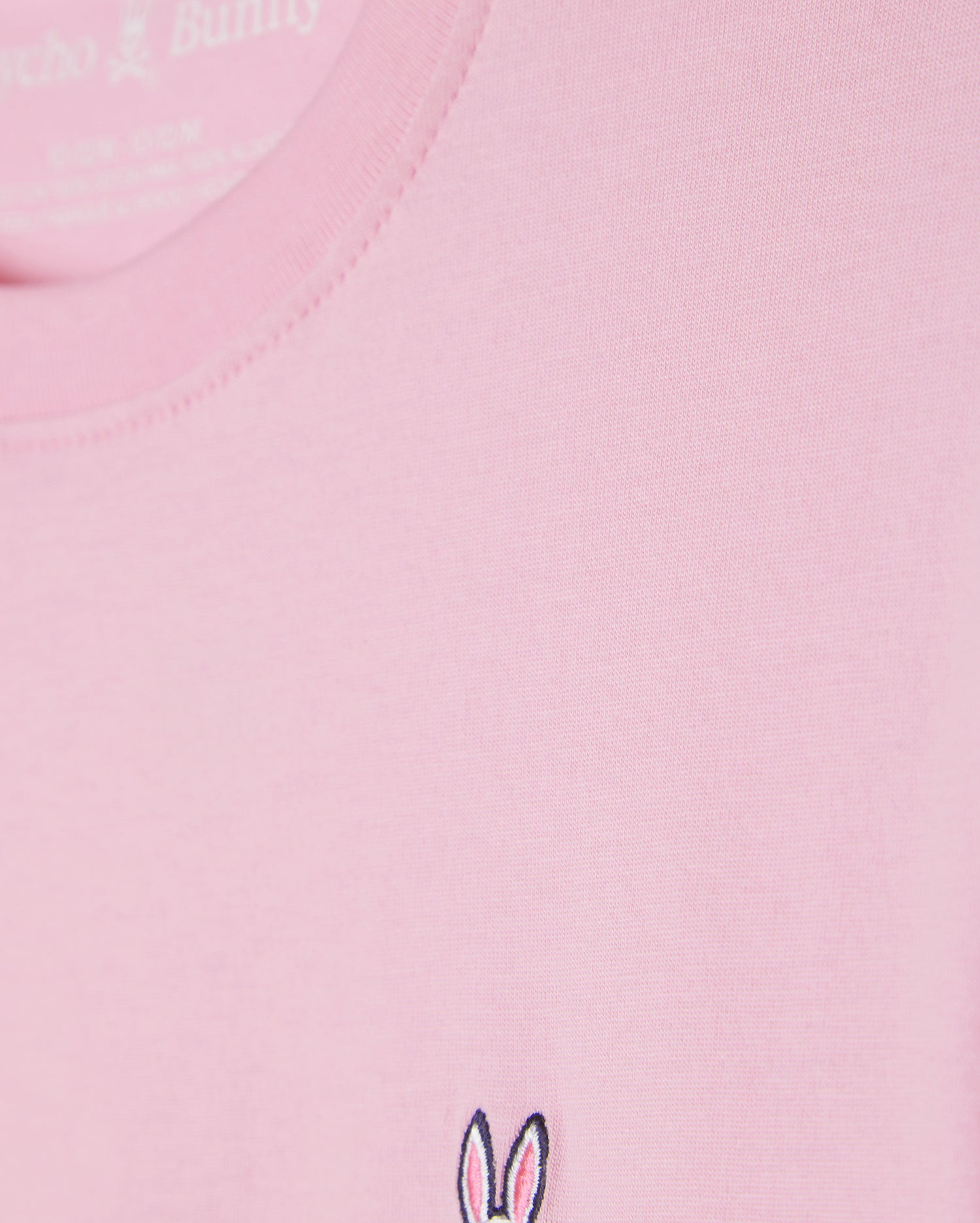 Close-up of the KIDS CLASSIC CREW NECK TEE - B0U014CRPC from Psycho Bunny, a light pink shirt made from 100% Pima cotton. It features a small embroidered bunny ears and skull logo near the hem, ideal for stylish back-to-school outfits.