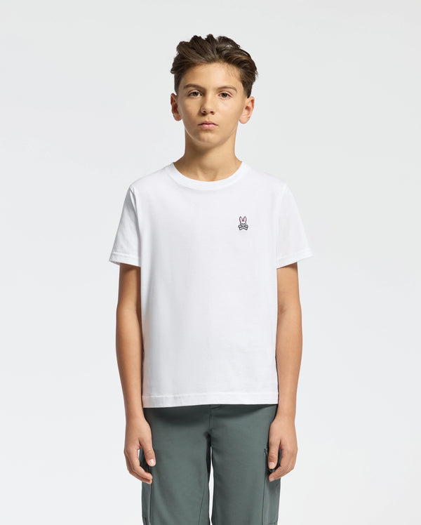 A young person with short dark hair, wearing a Psycho Bunny KIDS CLASSIC CREW NECK TEE - B0U014CRPC in crisp white 100% Pima cotton with the Classic Psycho Bunny embroidered logo, stands against a white background. They pair it with gray pants and maintain a neutral expression while facing forward.