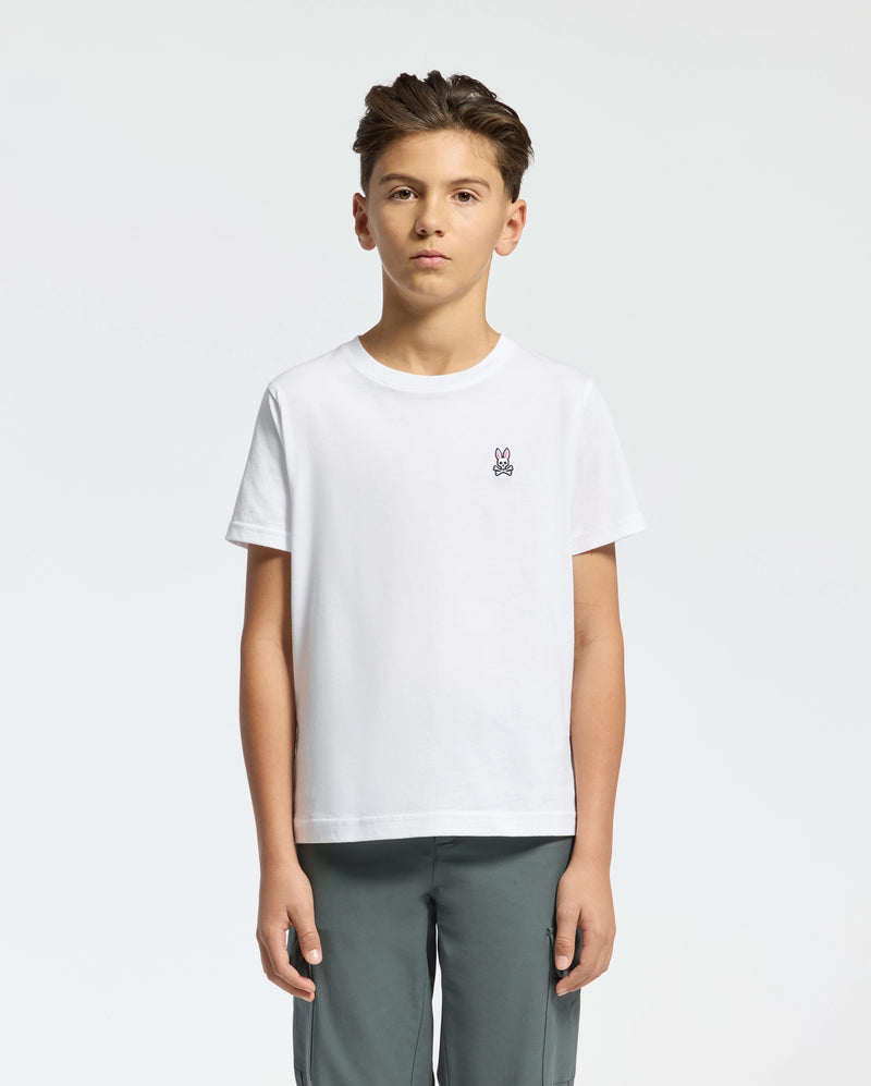 A young person with short dark hair, wearing a Psycho Bunny KIDS CLASSIC CREW NECK TEE - B0U014CRPC in crisp white 100% Pima cotton with the Classic Psycho Bunny embroidered logo, stands against a white background. They pair it with gray pants and maintain a neutral expression while facing forward.