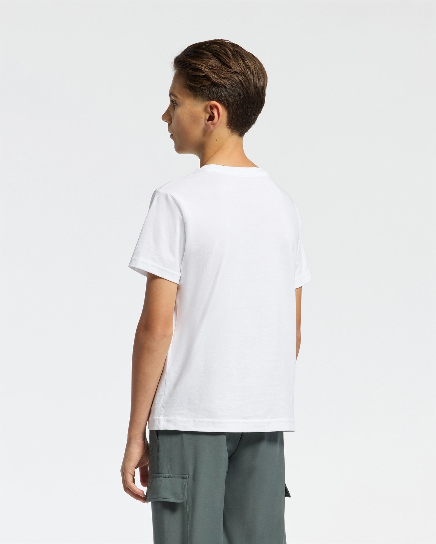 A young person with short brown hair stands sideways wearing Psycho Bunny's Kids Classic Crew Neck Tee in 100% Pima cotton and gray cargo pants. The plain white shirt showcases the classic embroidered Psycho Bunny logo on a neutral background.