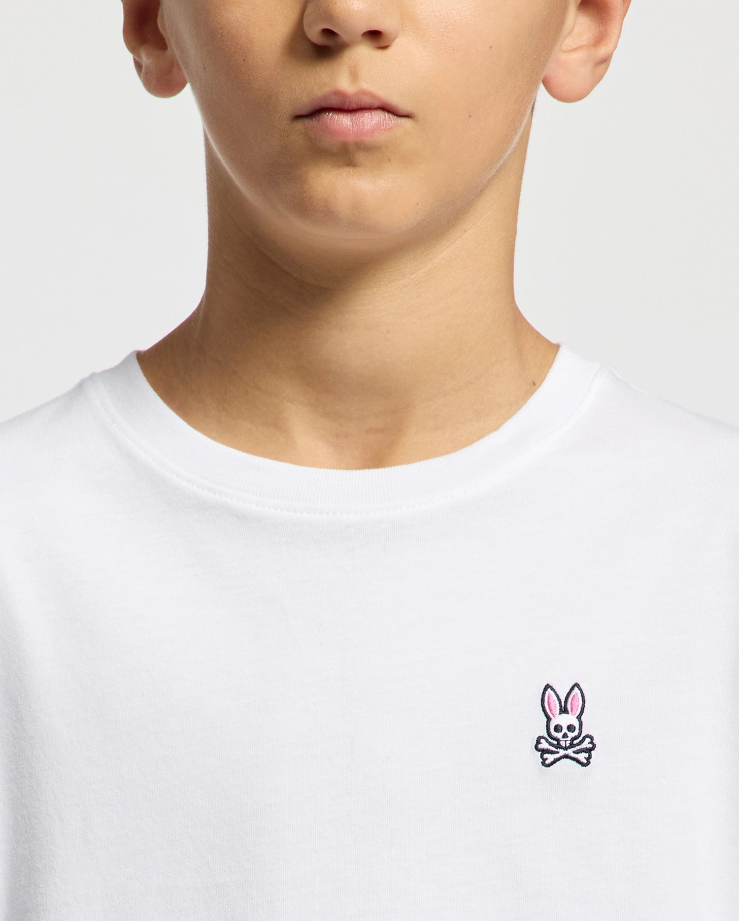 A person is seen wearing the Psycho Bunny Kids Classic Crew Neck Tee - B0U014CRPC, made from 100% Pima cotton, with a pink bunny over crossbones logo on the chest. Their lower face and upper torso are visible against a plain background.