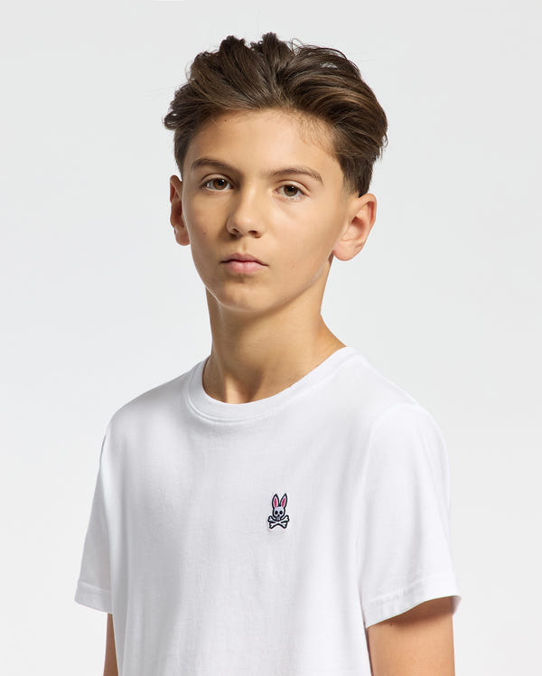 A boy with short brown hair wears the Psycho Bunny KIDS CLASSIC CREW NECK TEE (B0U014CRPC), featuring the Classic Psycho Bunny embroidered logo on his chest, perfectly complementing the plain white background. Made of 100% Pima cotton.