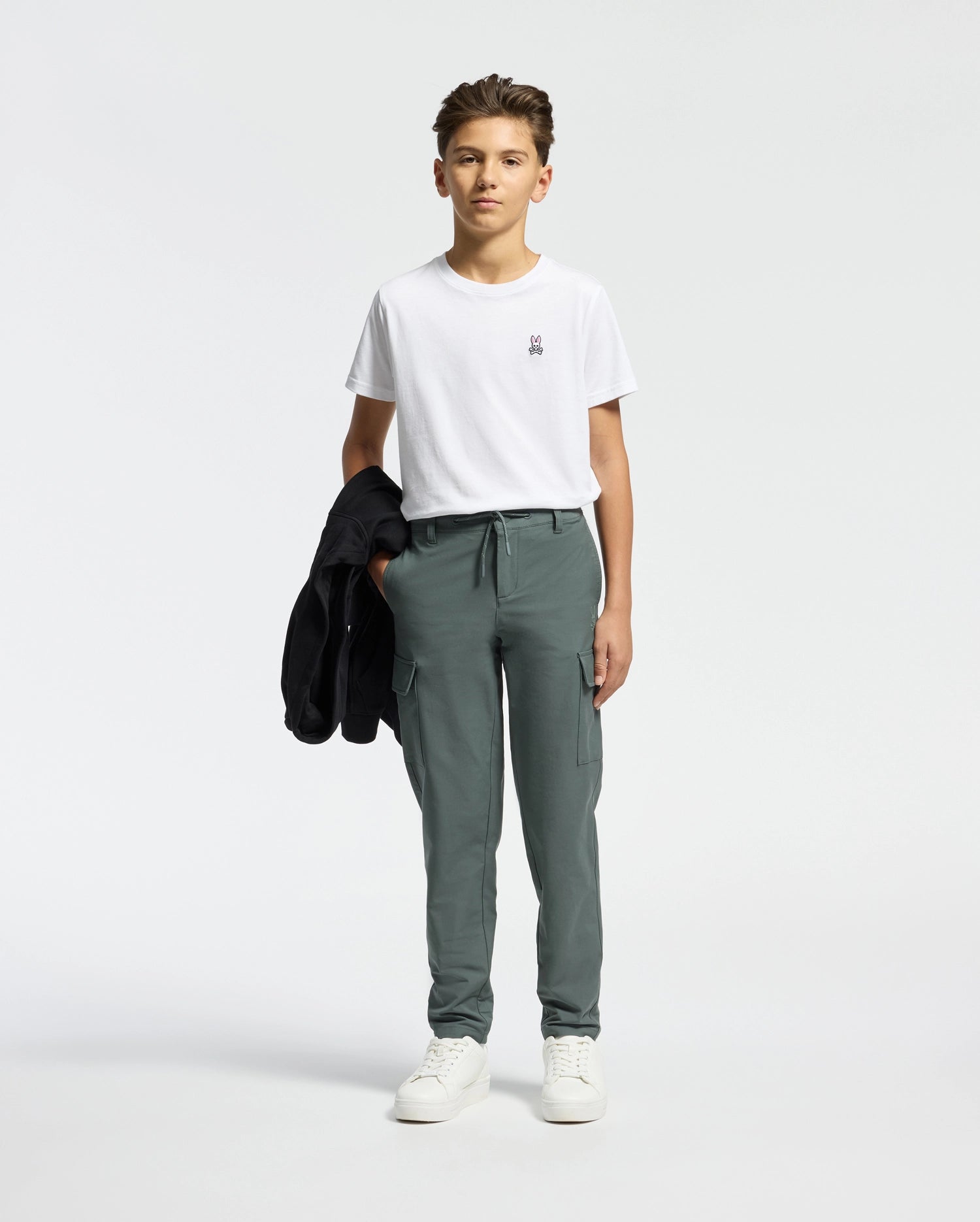 A young person stands against a plain background, wearing the Psycho Bunny KIDS CLASSIC CREW NECK TEE, green cargo pants, and white sneakers. They hold a black jacket on their shoulder, subtly nodding to the Classic Psycho Bunny logo.