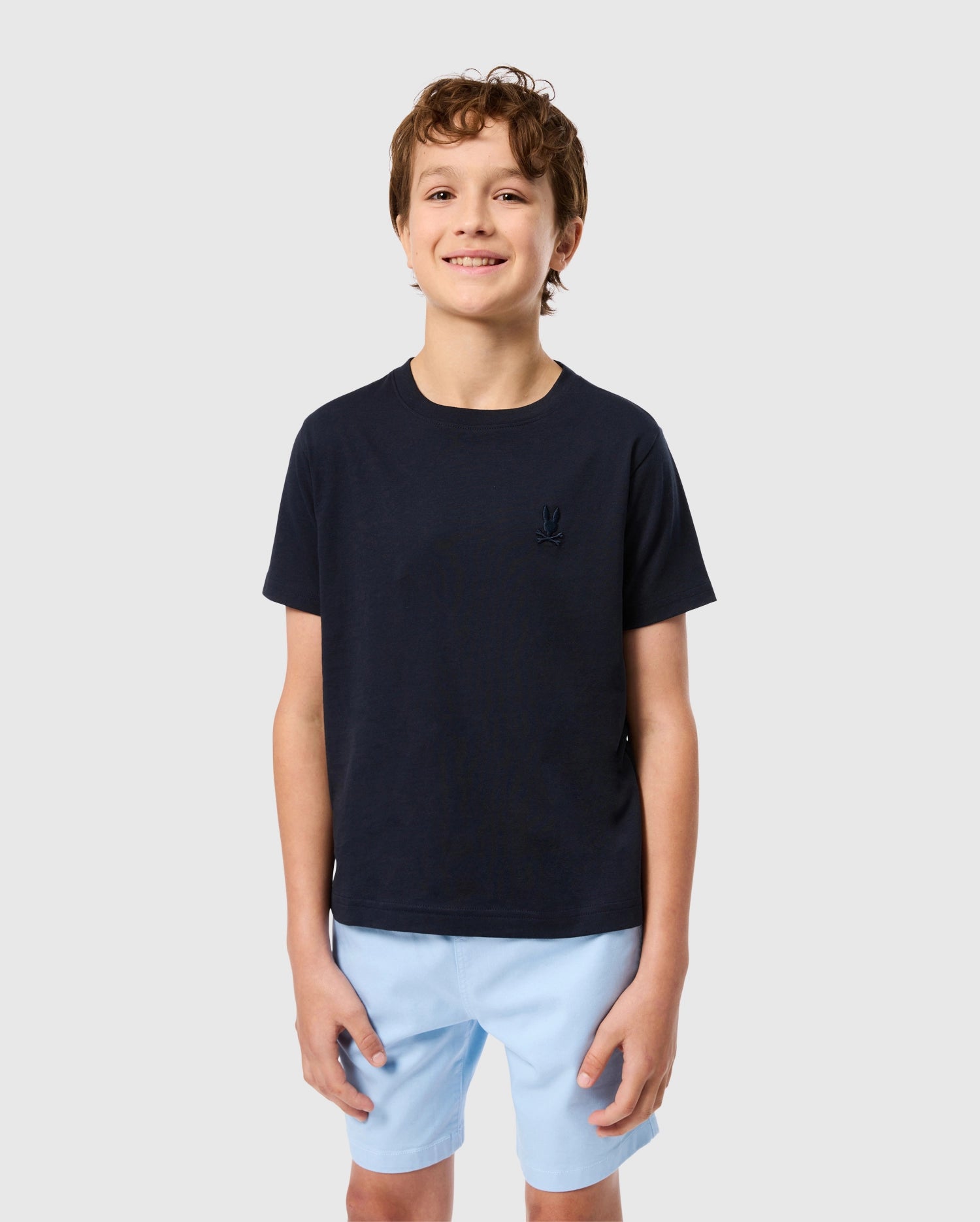 Against a plain gray background, a smiling boy wears the Psycho Bunny KIDS EMBROIDERED CLASSIC TEE in dark navy, crafted from soft Peruvian Pima cotton, with an embroidered chest bunny emblem, paired with light blue shorts.