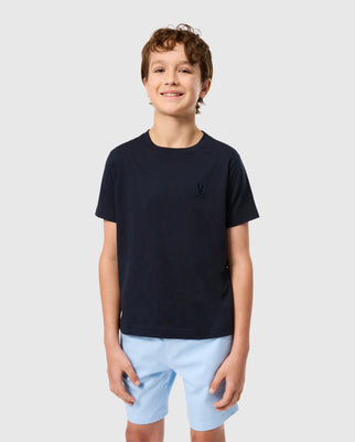 Against a plain gray background, a smiling boy wears the Psycho Bunny KIDS EMBROIDERED CLASSIC TEE in dark navy, crafted from soft Peruvian Pima cotton, with an embroidered chest bunny emblem, paired with light blue shorts.