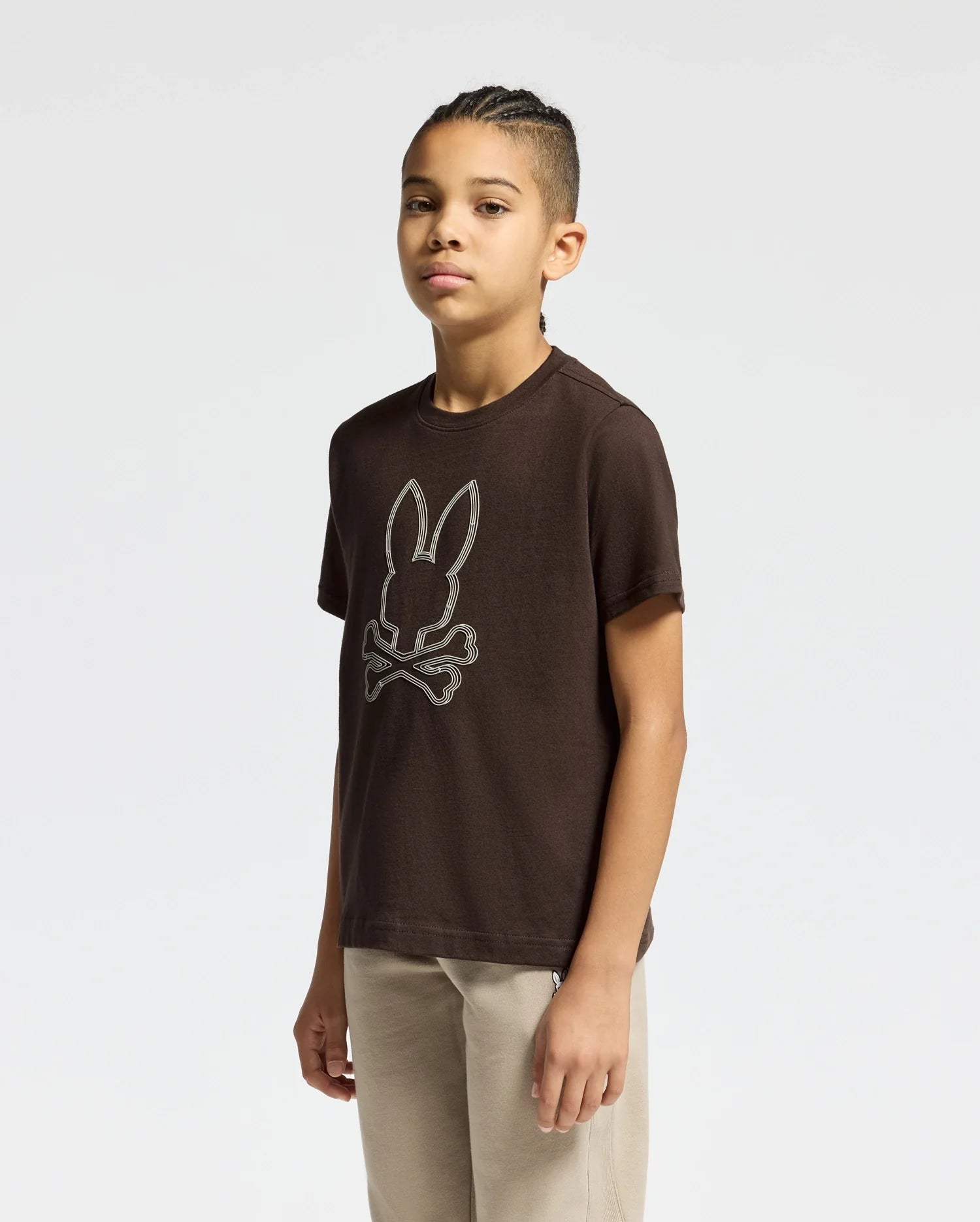 A young individual with braided hair is seen wearing a Psycho Bunny KIDS MONARC FRONT BUNNY GRAPHIC TEE in brown, featuring a rabbit skull and crossbones design. This unique Pima cotton graphic tee is paired with beige pants against a plain white backdrop.