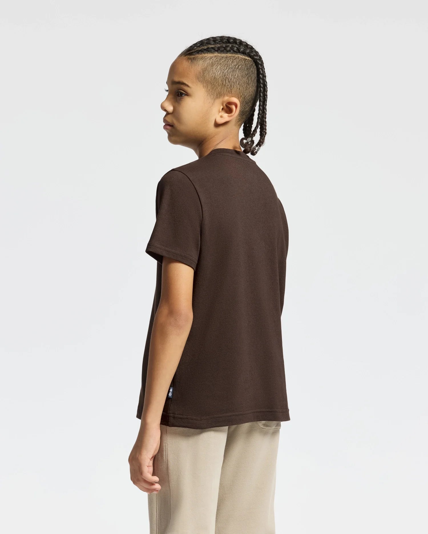 A child with long braided hair is standing and looking to the left, dressed in a dark brown Psycho Bunny KIDS MONARC FRONT BUNNY GRAPHIC TEE (B0U211E200) made from soft Pima cotton, paired with light beige pants. The background is plain and light gray.