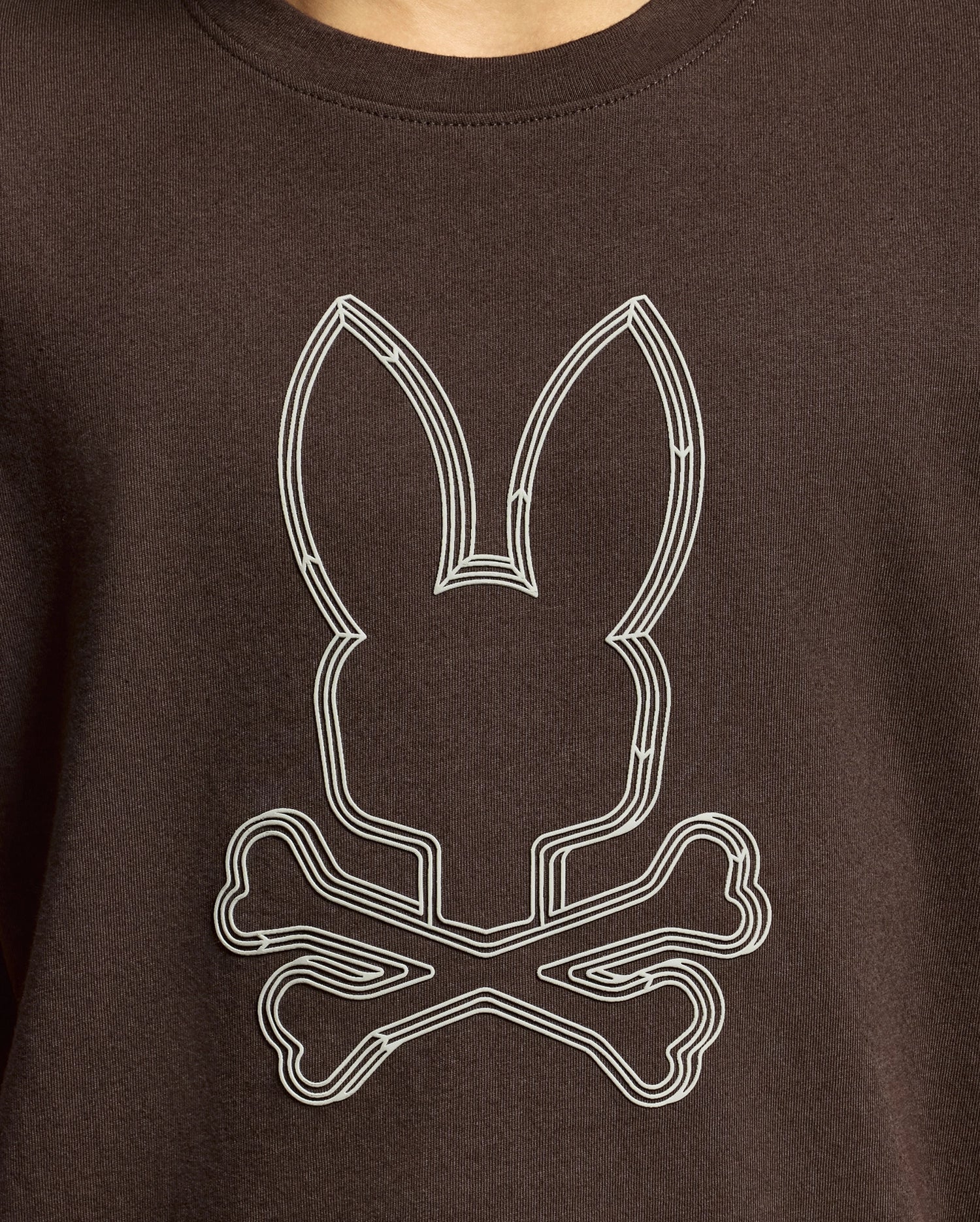 A close-up of the Psycho Bunny KIDS MONARC FRONT BUNNY GRAPHIC TEE - B0U211E200, made from soft Pima cotton, features a white geometric design of a rabbit's head with ears placed over two crossed bones on its rich dark brown fabric.