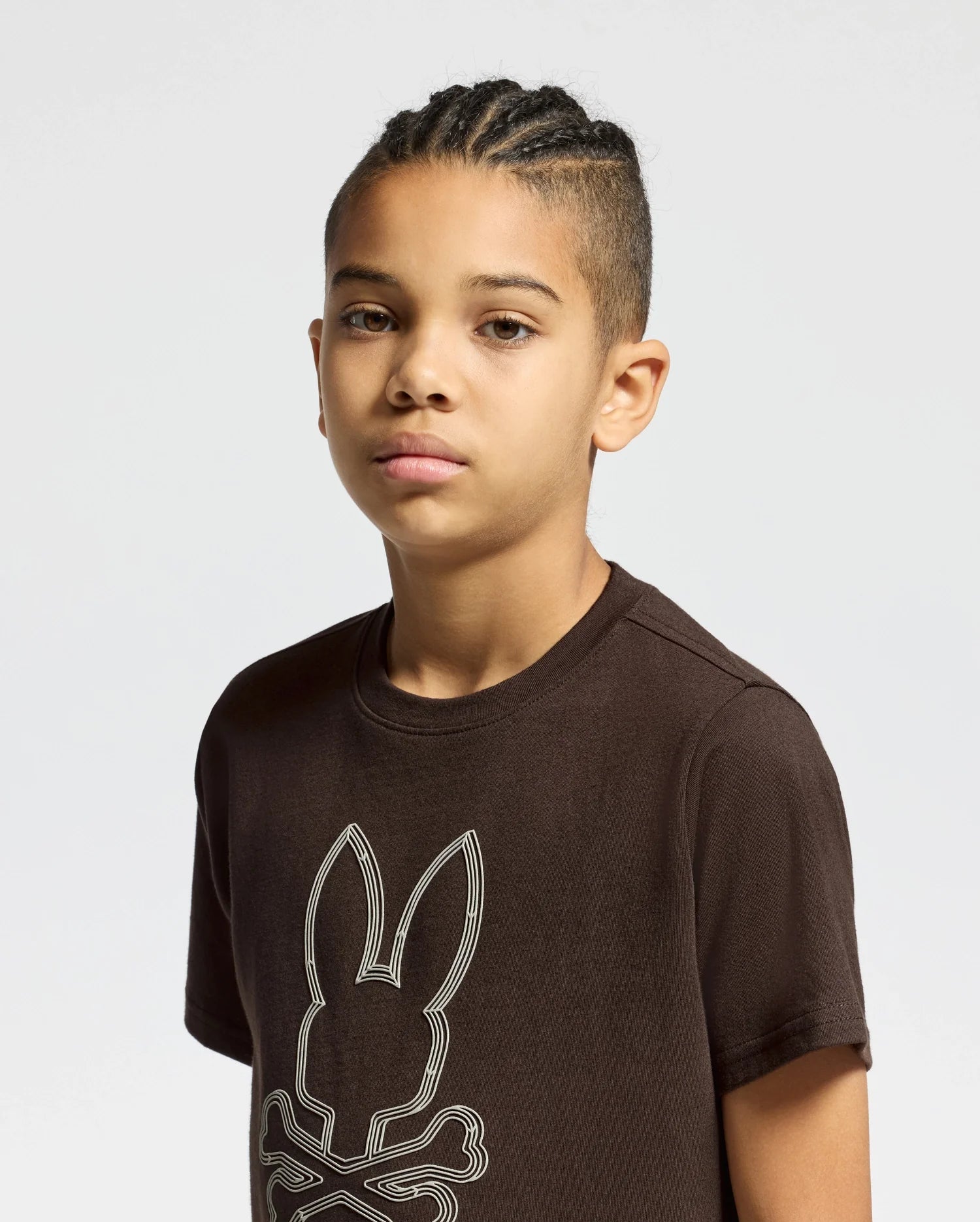 A young person with short braided hair is wearing a Kids Monarc Front Bunny Graphic Tee from Psycho Bunny, crafted from soft Pima cotton. They are looking directly at the camera against a plain, light-colored background.
