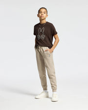 A young person stands in a relaxed pose, wearing a Psycho Bunny KIDS MONARC FRONT BUNNY GRAPHIC TEE - B0U211E200 crafted from soft Pima cotton, paired with beige joggers. White sneakers complete the casual look against a plain white background.