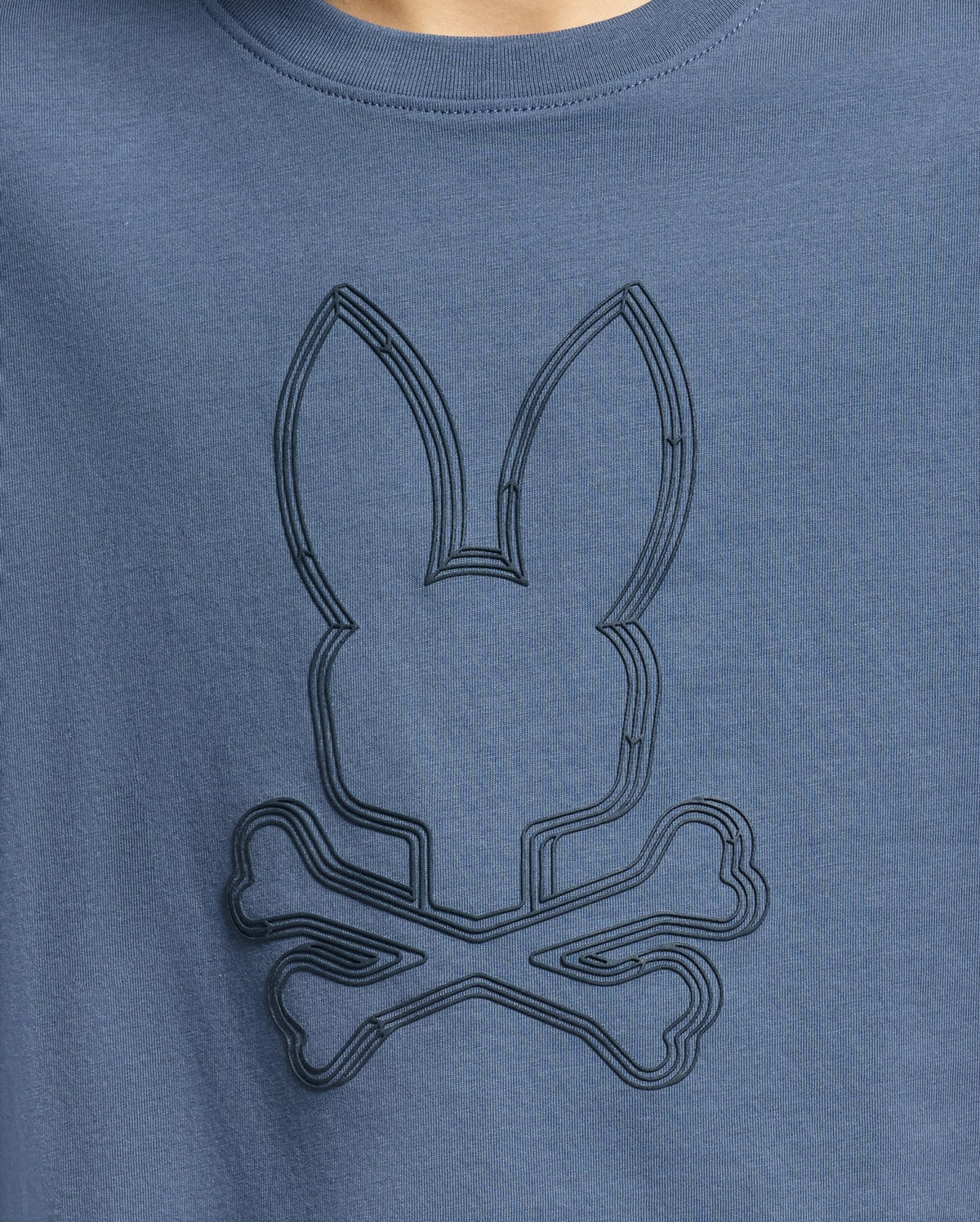 A close-up of the Kids Monarc Front Bunny Graphic Tee by Psycho Bunny, product code B0U211E200, in blue reveals a stylized, raised-printed bunny head above crossed bones, emulating a skull and crossbones motif. The design is crafted on premium Pima cotton in a slightly darker shade for subtle contrast.