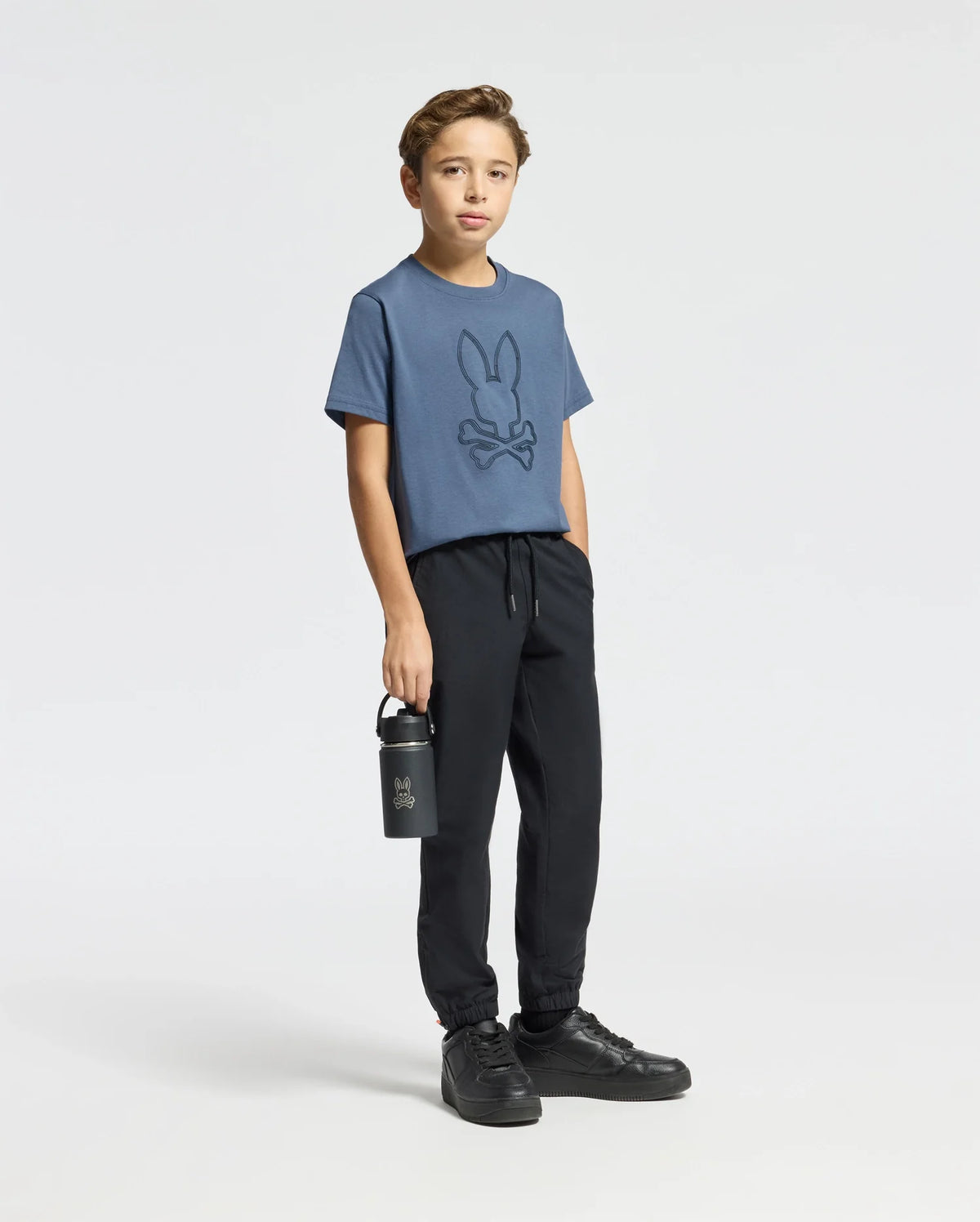 A young boy stands against a simple background wearing a blue KIDS MONARC FRONT BUNNY GRAPHIC TEE - B0U211E200 by Psycho Bunny, crafted from soft Pima cotton. He pairs it with black joggers and sneakers while holding a black water bottle in his left hand.