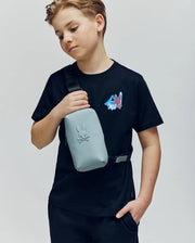 A boy in a Psycho Bunny KIDS MONTREAL TEE (B0U381F200)—black with a small colorful graphic—pairs it with a light gray crossbody bag, standing with one hand in his pocket. Made from 100% Pima cotton, it channels cool Montreal style.