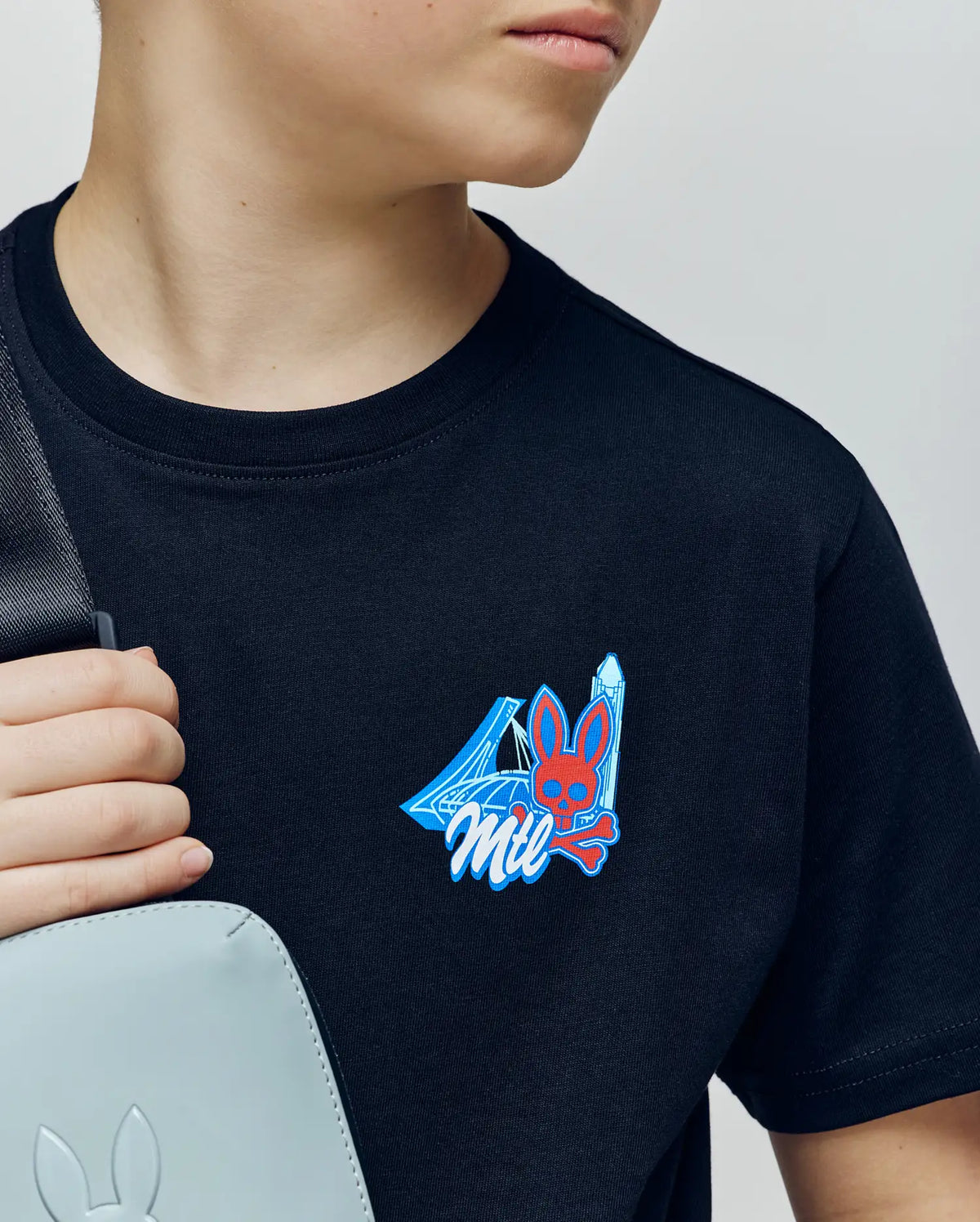 A child wears the Psycho Bunny KIDS MONTREAL TEE (B0U381F200), a black 100% Pima cotton shirt with a blue and red bunny skull, crystals, and "Mil" graphic, paired with a light gray bag featuring a subtle bunny logo.