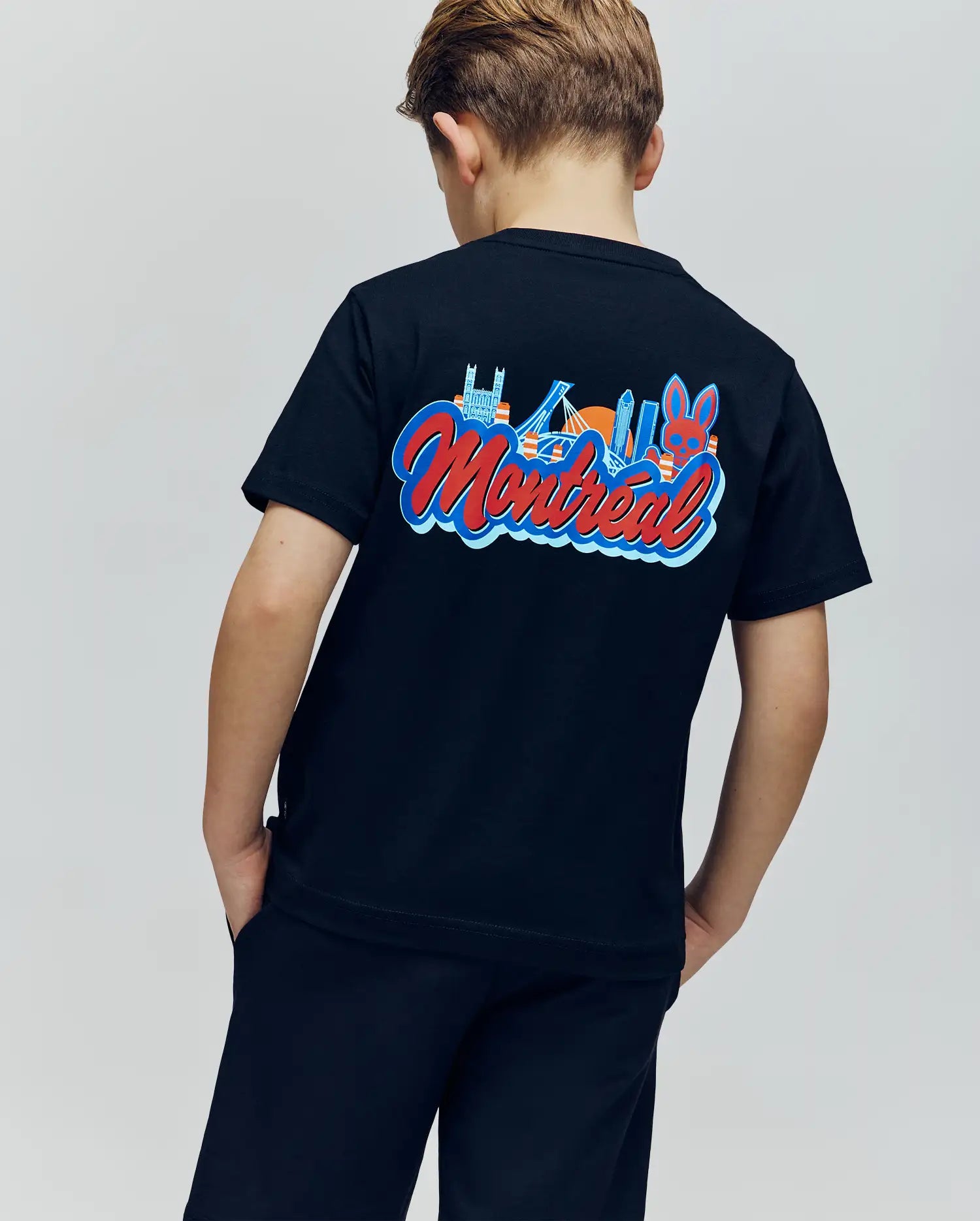 A child faces away wearing the Psycho Bunny KIDS MONTREAL TEE (B0U381F200), a black 100% Pima cotton T-shirt with colorful Montreal landmarks and "Montreal" on the back, paired with black shorts.