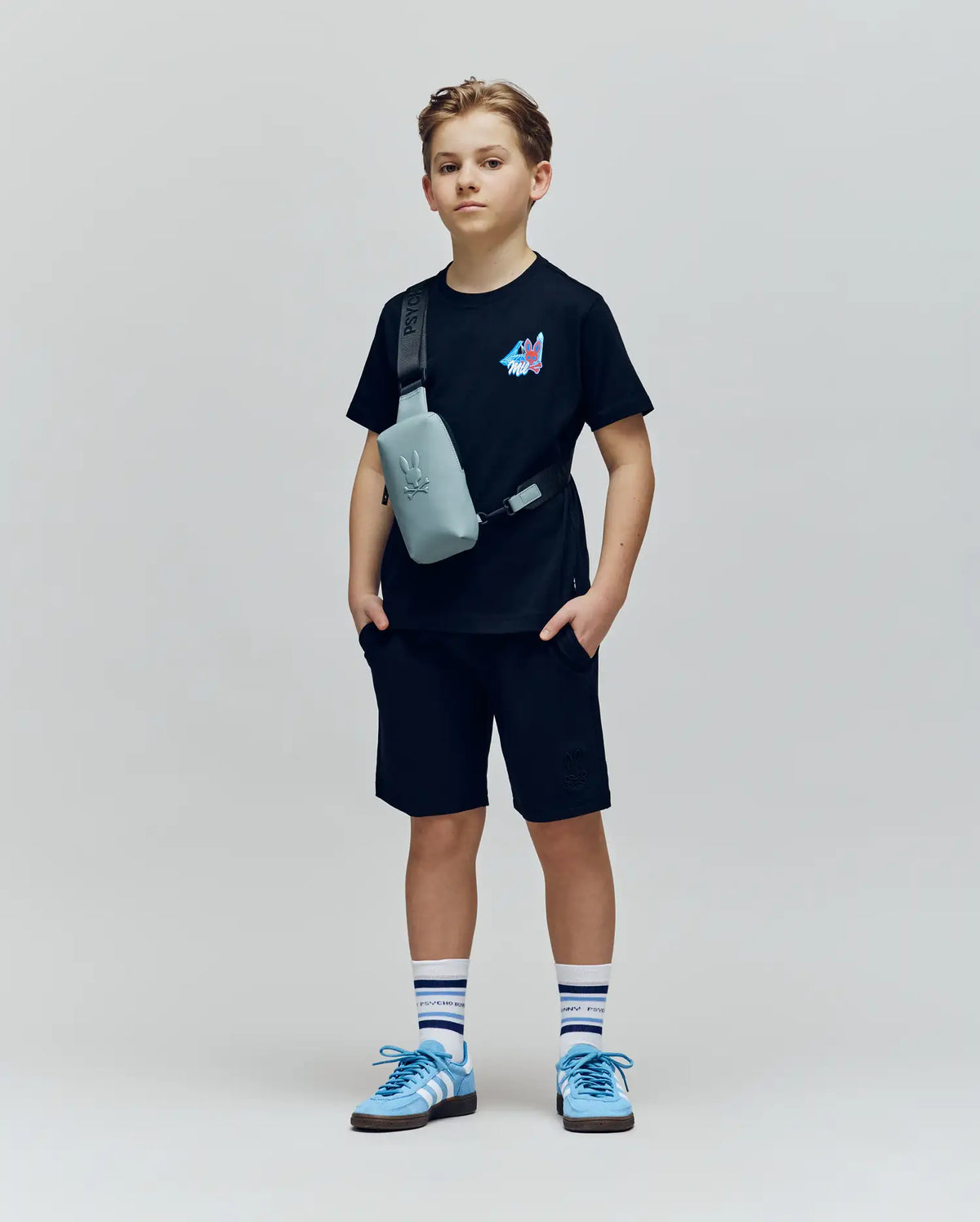 A young boy stands against a plain background wearing the Psycho Bunny KIDS MONTREAL TEE (B0U381F200), black shorts, blue sneakers, striped socks, and a gray crossbody bag. He looks straight ahead with hands in his pockets.
