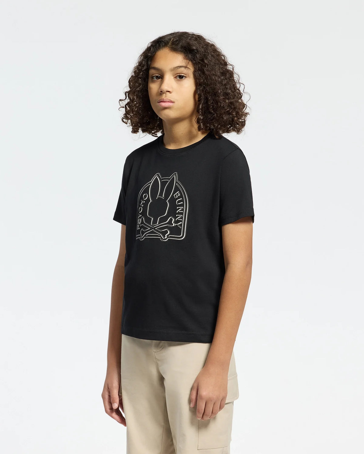 Someone with curly hair is wearing a Psycho Bunny Melbourne graphic tee made from eco-friendly Peruvian Pima cotton. The black tee has a bunny with a bow tie design and "Bunny" text. They wear it with light-colored pants against a plain, light background.