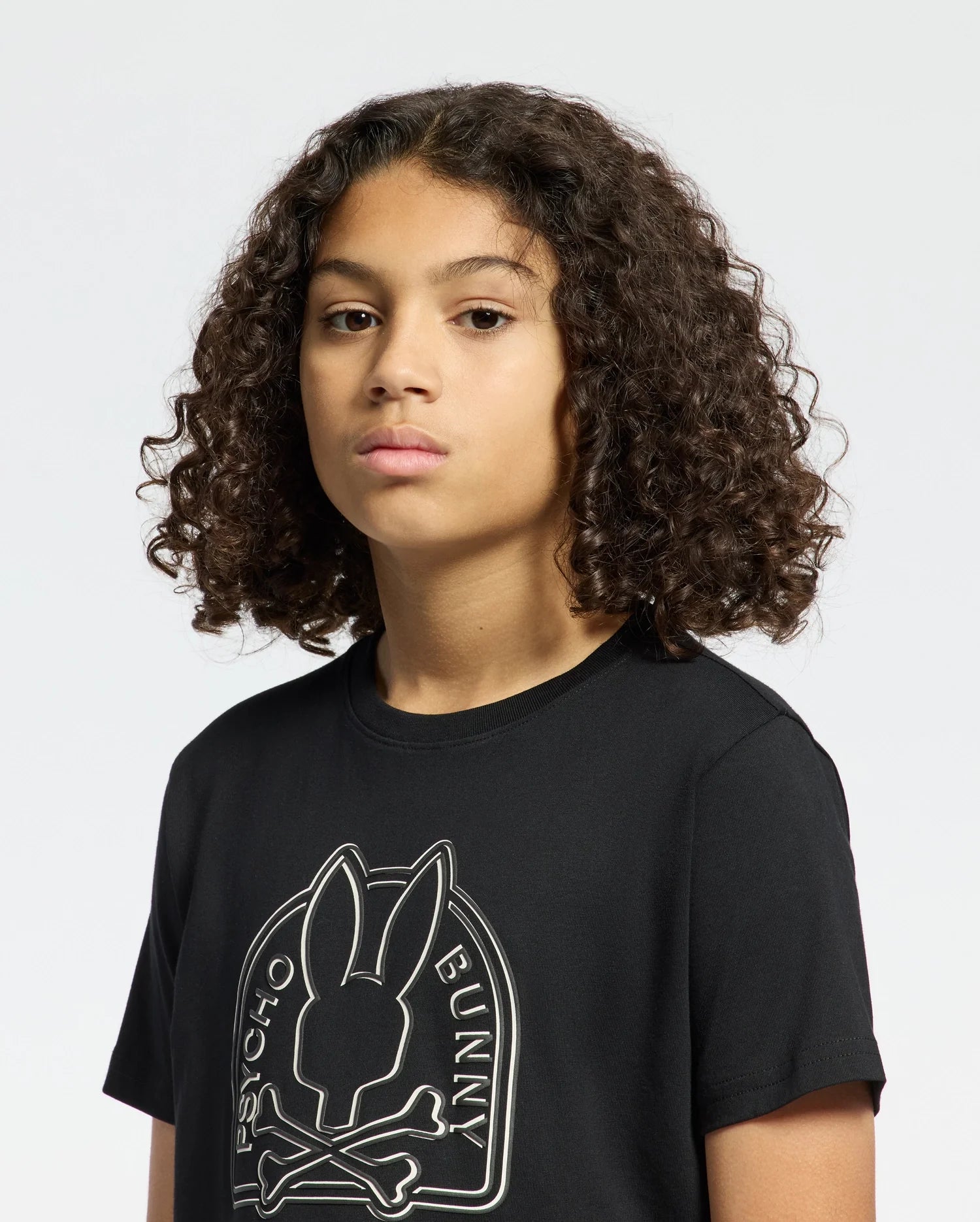 A person with curly hair wears a black KIDS MELBOURNE GRAPHIC TEE (B0U410F200) by Psycho Bunny, crafted from eco-friendly Peruvian Pima cotton and featuring a bunny logo with bones. They look ahead neutrally against a plain backdrop.