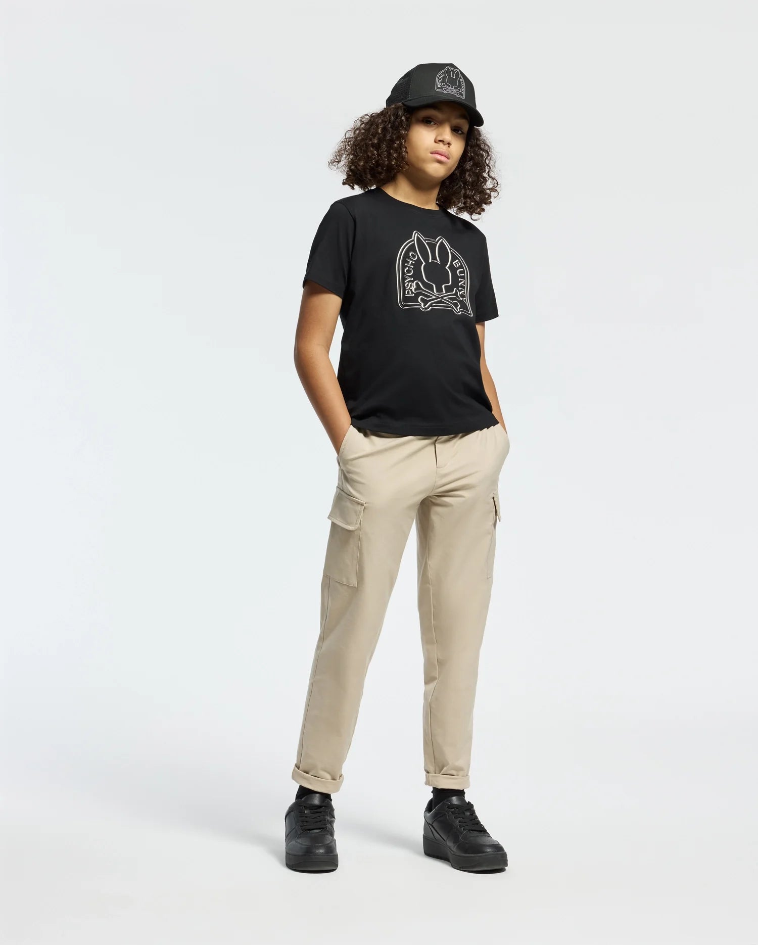 A person with curly hair is wearing a black cap, a Psycho Bunny KIDS MELBOURNE GRAPHIC TEE featuring line art, beige cargo pants, and black sneakers. They're standing casually against a plain white background with one hand in their pocket.