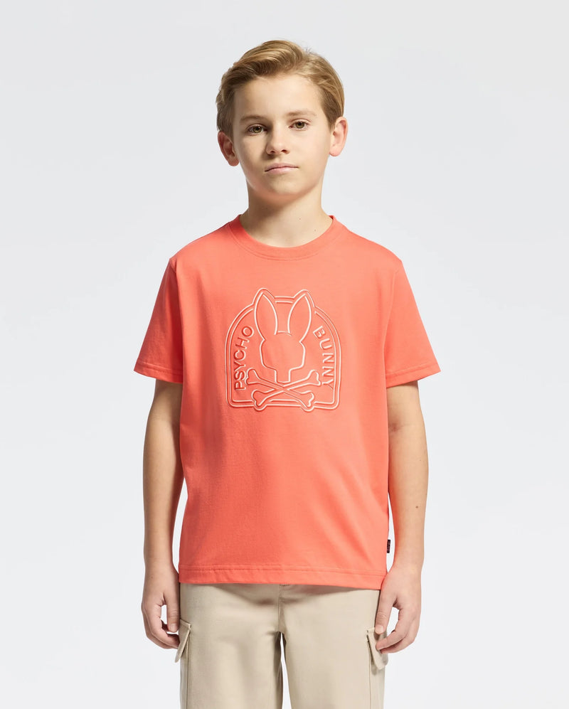 A young boy with light brown hair is wearing a Psycho Bunny KIDS MELBOURNE GRAPHIC TEE (B0U410F200) in orange, featuring a bunny and crossbones design. It's crafted from soft Pima cotton and paired with beige cargo pants against a plain white background.
