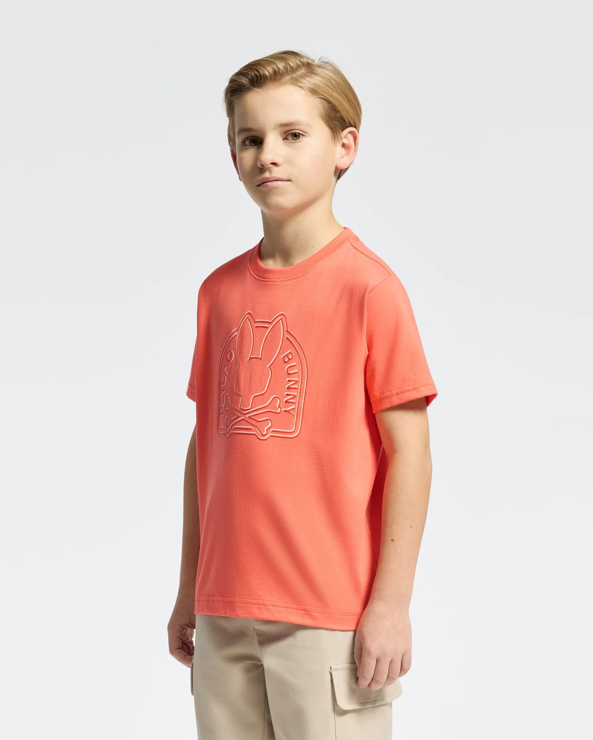 A boy in a colorful Psycho Bunny KIDS MELBOURNE GRAPHIC TEE (B0U410F200) with a bunny design stands against a white background. His left arm is bent slightly, and he's paired the shirt with comfy beige pants, creating a charming casual style.