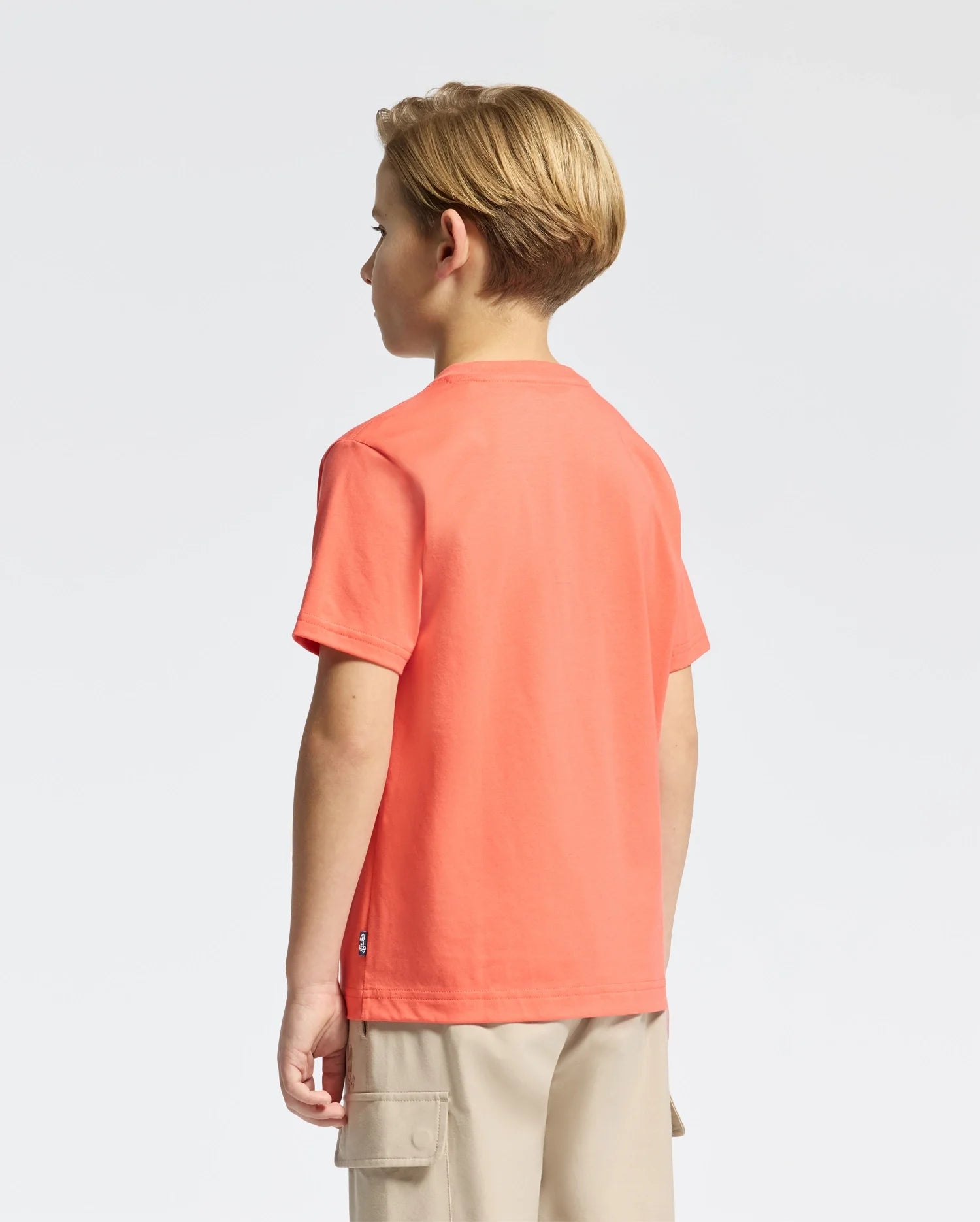 A young blonde-haired boy faces away, wearing a Psycho Bunny KIDS MELBOURNE GRAPHIC TEE in bright orange, made from soft Pima cotton and featuring a charming bunny design. He pairs it with beige cargo shorts against a plain white background.