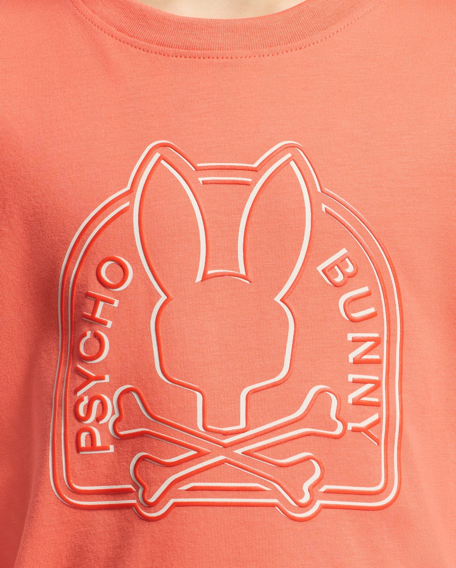 Close-up of a coral-colored KIDS MELBOURNE GRAPHIC TEE by Psycho Bunny, showcasing a raised Bunny graphic with a stylized bunny head above crossed bones and "Psycho Bunny" written along the sides.