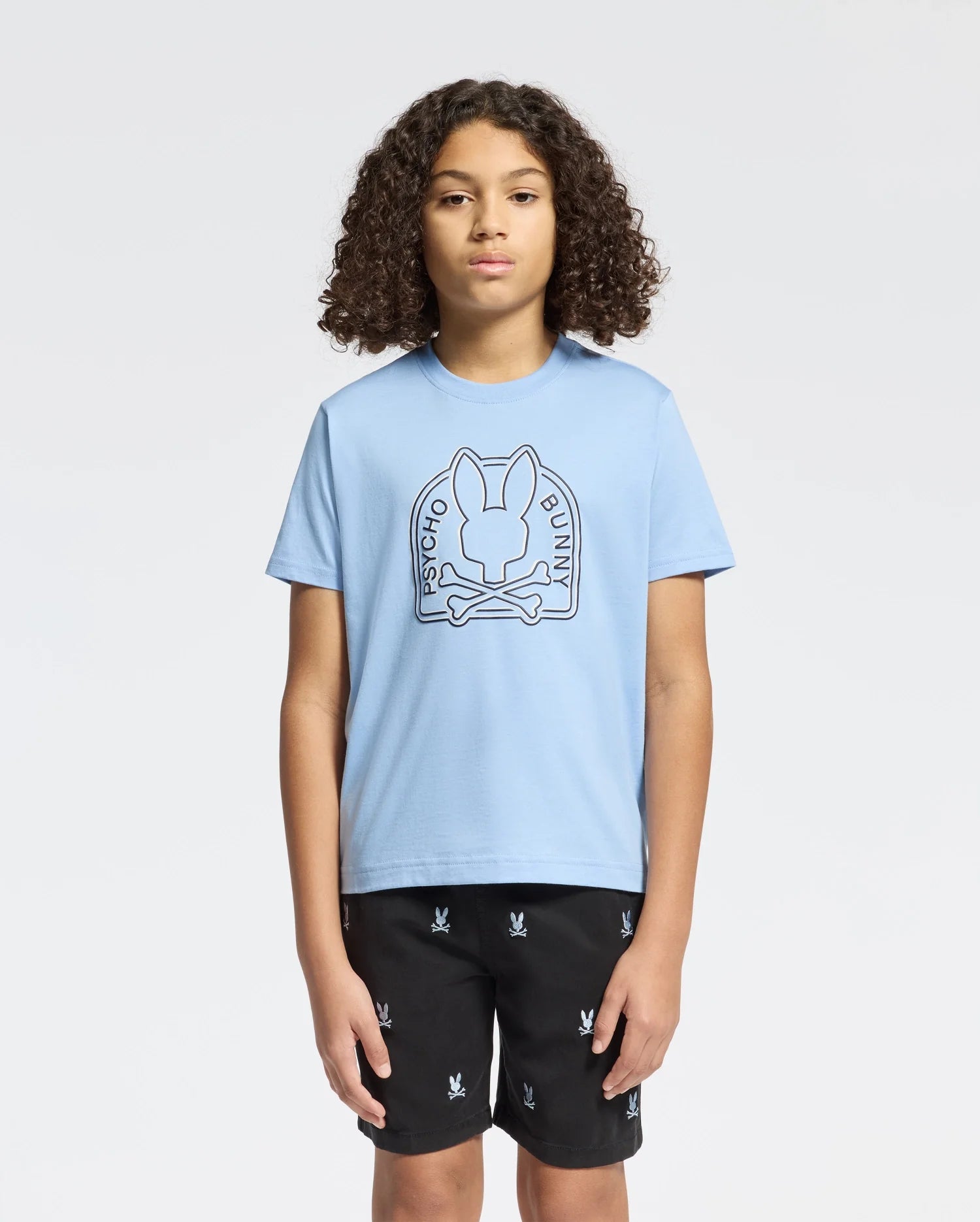 A person with curly hair wears an eco-friendly light blue KIDS MELBOURNE GRAPHIC TEE (B0U410F200) by Psycho Bunny, featuring a bunny and crossbones design. They're also in black shorts with small motifs against a plain light gray backdrop.